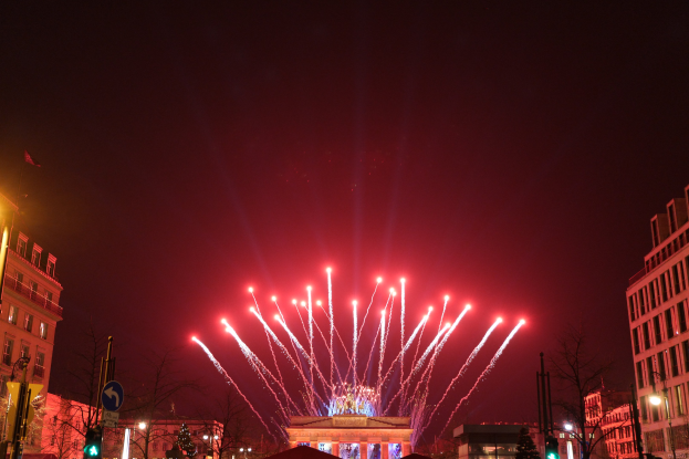Eine belebte nächtliche Stadtstraße in Berlin, Deutschland, mit dem Brandenburger Tor in der Mitte, beleuchtet von Gebäudelichtern, Bäumen, Pfosten, Verkehrsampeln, Schildern und Flaggen, und Feuerwerk, das den Himmel über dem Tor erhellt.