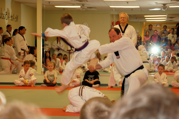 Zwei Personen kämpfen in einem Raum mit Kindern auf dem Boden, eine Gruppe von Menschen, eine Kamera, eine Uhr, eine Wand, ein Dach, Deckenleuchten, ein Ventilator, eine Säule und eine Wand im Hintergrund.