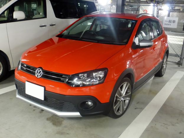 Ein oranges Volkswagen Polo Cross 1.6 TDI Highline DSG, das in einer Garage neben einem weißen Transporter geparkt ist, mit einem Metallzaun, einer Tafel mit Text, Lampen und einer Decke im Hintergrund.