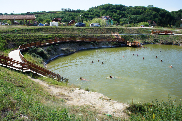 Gruppe von Menschen, die in einem Gewässer mit üppiger Vegetation, einer Brücke mit Treppen, Hütten, Fahrzeugen, Pfählen und einem klaren blauen Himmel im Hintergrund schwimmen.