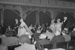 Ein Schwarz-Weiß-Foto einer Hochzeitszeremonie in einem großen Raum mit vielen sitzenden und stehenden Gästen, einige halten Blumensträuße, und Tische mit Tassen und anderen Gegenständen, mit Vorhängen und Deckenlampen im Hintergrund.