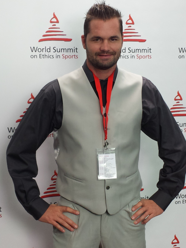 Ein Mann im Anzug, der eine schwarze Hemd, graue Hose und einen Ausweis trägt, steht vor einem Banner, auf dem 'World Summit on Ethics in Sports' steht.