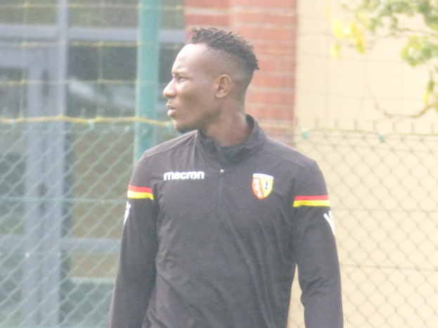 Ein Mann in einer schwarzen Jacke steht auf einem Fussballfeld umgeben von einem Zaun und einem Baum und sieht konzentriert aus während eines Trainings.