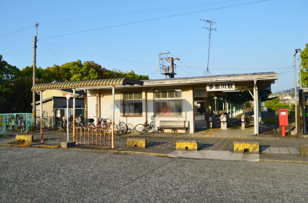Ein Bahnhof mit Fahrrädern davor, eine Straße unten und Häuser, Bäume, Pfähle, Drähte und einen klaren blauen Himmel im Hintergrund.