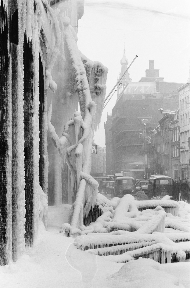 Ein Schwarz-Weiß-Foto einer verschneiten und vereisten Stadtstraße mit Gebäuden, Fahrzeugen, Menschen, Pfählen und einem Himmel im Hintergrund.