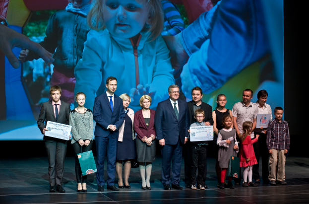Eine Gruppe von Menschen auf einer Bühne, einige halten Taschen und Urkunden in den Händen, im Hintergrund ein Bildschirm mit dem Bild eines Kindes und anderer Menschen.