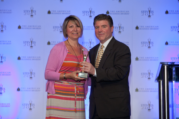 Zwei Personen mit einem Pokal, wobei eine Person einen schwarzen Blazer und ein weißes Hemd trägt und ein Banner im Hintergrund zu sehen ist.