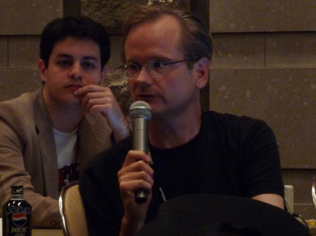 Zwei Personen sitzen auf Stühlen, eine in einem Blazer und die andere in einem schwarzen T-Shirt mit einem Mikrofon; eine Flasche mit schwarzem Deckel und einem Markennamen ist links.