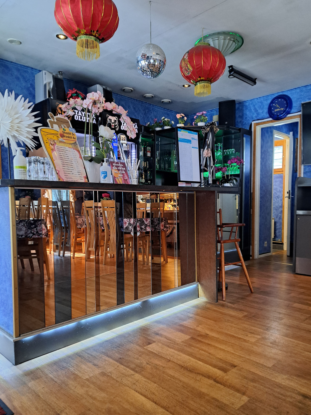 Das Innere eines Restaurants mit blauen Wänden und roten Laternen, die von der Decke hängen, mit Tischen und Stühlen, einem zentralen Schreibtisch mit Schränken, Tafeln mit Text, Blumenvasen, Dekorationsgegenständen, einer Tür und einer Wanduhr.