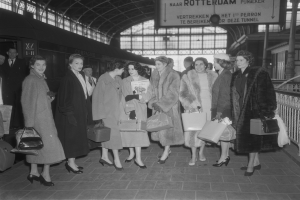 Schwarz-weißes Bild von Frauen, die an einem Bahnhof stehen und Taschen halten, mit einem Zug auf der linken Seite und Geländern und einer Tafel auf der rechten Seite.
