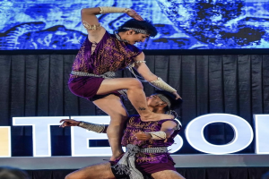 Zwei Performer in purpurfarbenen Kleidern führen Akrobatik auf der Bühne vor einem Publikum aus, mit einem schwarzen Vorhang und einem Bildschirm im Hintergrund.