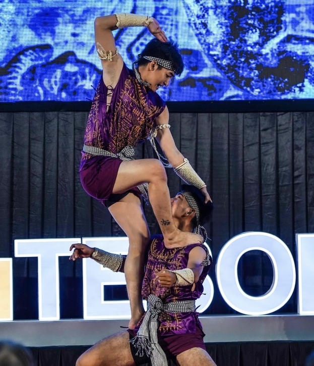 Zwei Performer in purpurfarbenen Kleidern führen Akrobatik auf der Bühne vor einem Publikum aus, mit einem schwarzen Vorhang und einem Bildschirm im Hintergrund.