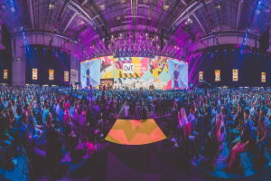 Eine große Menschenmenge auf Stühlen sitzend, die einer hell erleuchteten Bühne mit mehreren stehenden Individuen zugewandt sind, umgeben von Bildschirmen.