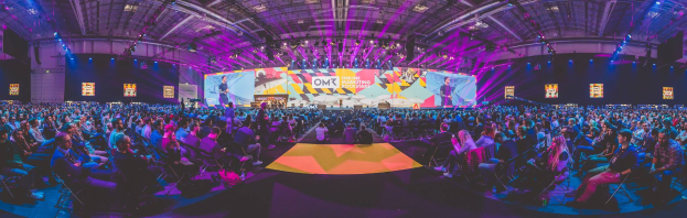 Eine große Menschenmenge auf Stühlen sitzend, die einer hell erleuchteten Bühne mit mehreren stehenden Individuen zugewandt sind, umgeben von Bildschirmen.