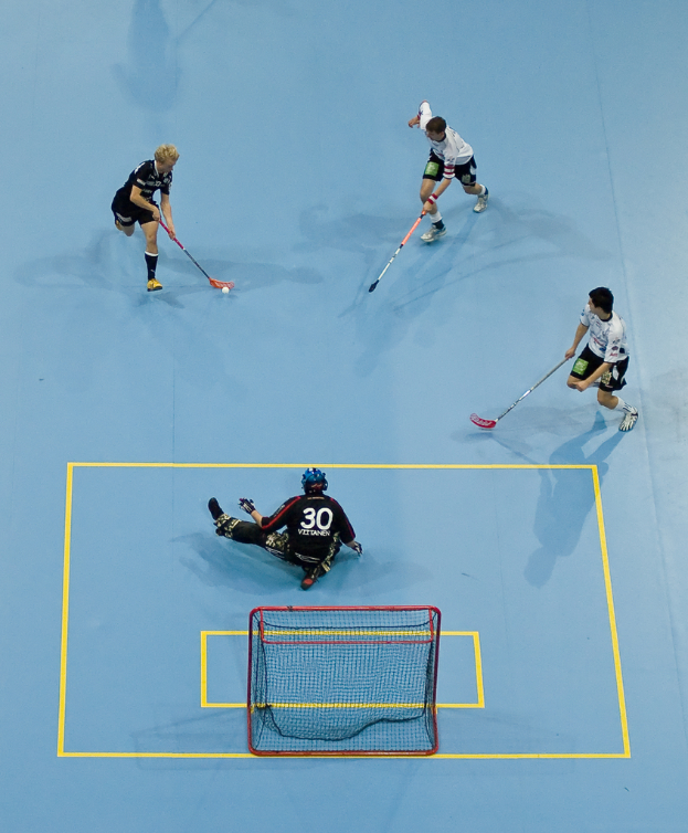 Drei Personen in Sportkleidung spielen Hockey, wobei eine Person rechts einen Ball mit einem Hockeystock schlägt und eine andere Person hinter einem Netz auf dem Boden sitzt.