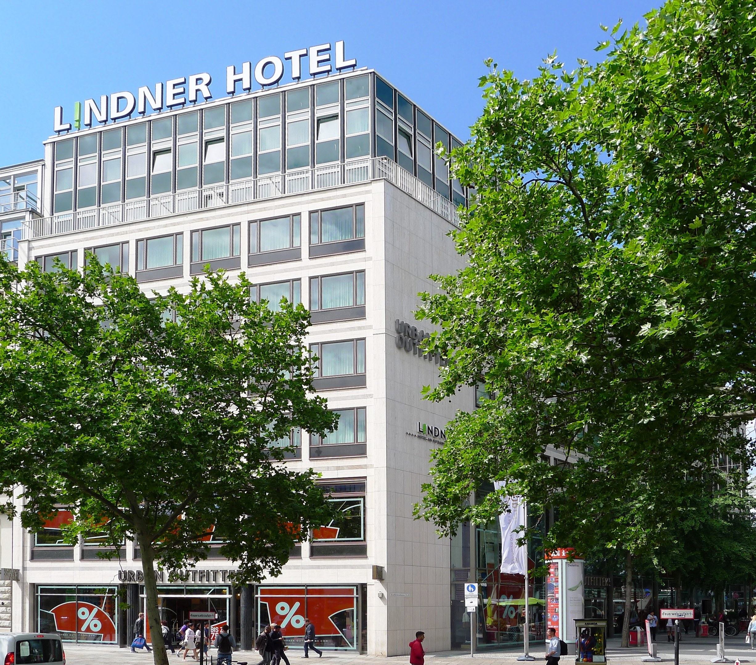 Großes Glasfrontgebäude des Lindner Hotels in Berlin, Deutschland, mit Text oben drauf, umgeben von Bäumen, Fahrzeugen und Fußgängern unter einem sichtbaren Himmel.