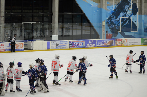 Group of young people playing ice hockey on an indoor rink, wearing helmets and holding sticks, with a person standing to the left and banners, pillars, and glass walls in the background.
