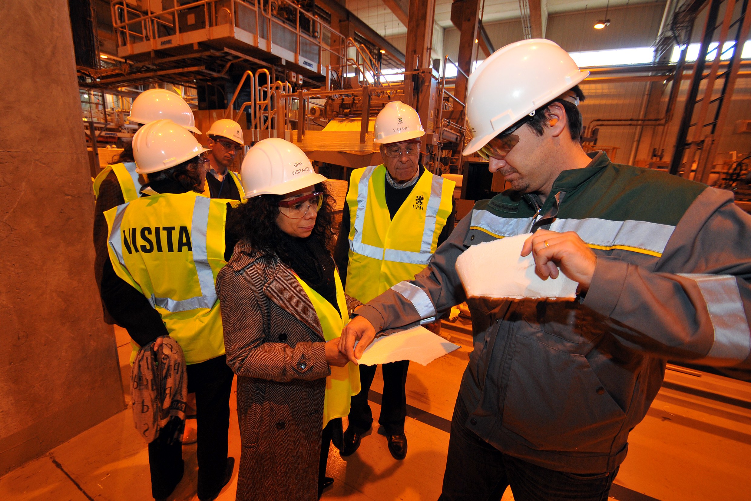 Gruppe von Menschen in Helmen und Sicherheitswesten in einer Fabrik, mit einem Mann, der Papiere und einen Stift hält, und einer Frau, die Papiere hält, in der Nähe von Metallstrukturen und einer Wand mit Deckenleuchten.