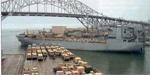Ein großes Schiff liegt nahe einer Brücke vor Anker, mit Fahrzeugen und Kisten auf dem Deck und Gebäuden im Hintergrund unter einem klaren blauen Himmel.