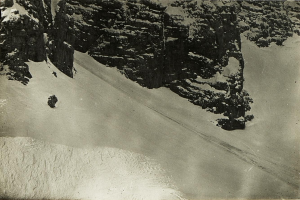 Ein Schwarz-Weiß-Foto eines Menschen, der eine schneebedeckte Bergabfahrt hinabfährt, umgeben von schneebedeckten Höhen, mit Text am unteren Rand.