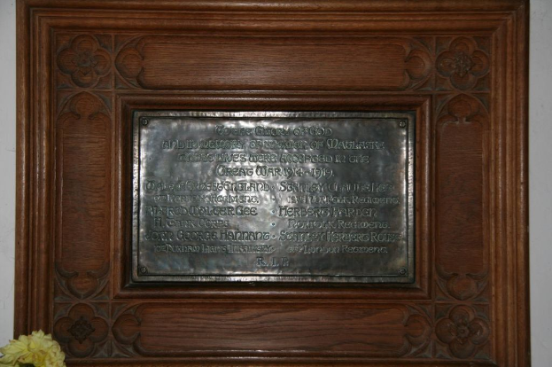 Eine Gedenktafel mit Text ist an einer braunen Wand oder einer Holztür angebracht, mit einem gelben Objekt, das einer Vase ähnelt, in der unteren linken Ecke.