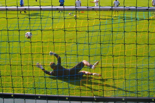 Ein Fussballer taucht ab, um einen Ball vor dem Tor zu halten, mit einem Netz im Vordergrund und einigen Menschen im Hintergrund, während mehrere Fussbälle auf dem Boden verstreut sind.