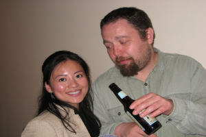 Ein Mann hält eine Flasche und schaut eine Frau an, die an einer Wand lehnt und lächelt.