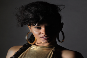 Eine Frau in einem goldenen Kleid und großen Kreolen steht selbstbewusst vor einer Wand, ihr glatter Bob und natürliches Make-up unterstreichen ihren entschlossenen Gesichtsausdruck, während sie direkt in die Kamera blickt.