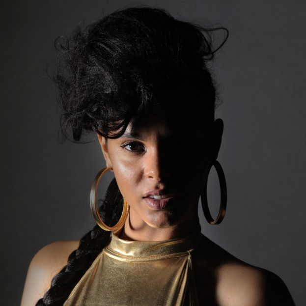 Eine Frau in einem goldenen Kleid und großen Kreolen steht selbstbewusst vor einer Wand, ihr glatter Bob und natürliches Make-up unterstreichen ihren entschlossenen Gesichtsausdruck, während sie direkt in die Kamera blickt.