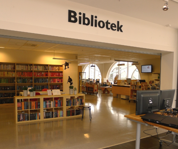 Mehrere Holzregale voller Bücher, mit Monitoren auf der rechten Seite eines Tisches und dem Wort 'BIBLIOTECA' oben angezeigt.