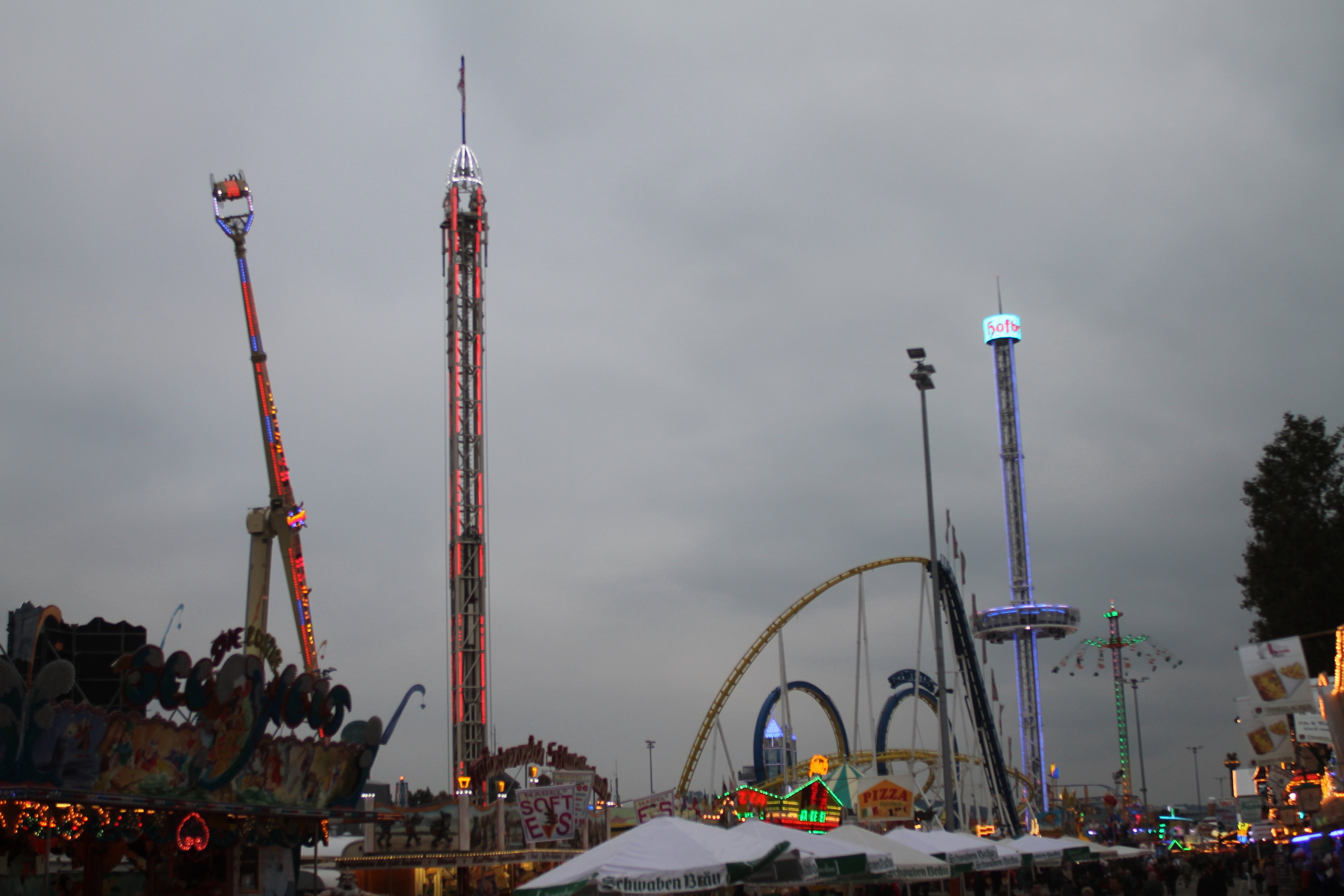 Eine Menschenmenge versammelt sich um einen hell erleuchteten Fasching bei Nacht, mit beleuchteten Fahrgeschäften, Pfählen und Bäumen unter einem bewölkten Himmel.