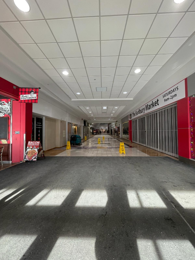 Das Innere eines Einkaufszentrums mit Deckenlampen, einem Stuhl und einer Tafel, auf der "Sydbury Market - Before and After" steht, und verstreuten Gegenständen auf dem Boden.