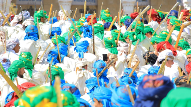 Eine große Gruppe von Menschen in bunten Kostümen marschiert auf einer Straße, jeder hält einen Stock, während eines Festivals mit einem Gebäude im Hintergrund.
