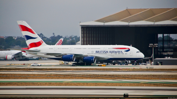 Ein Flugzeug auf einer Rollbahn mit Straßenlaternen rechts daneben, ein Auto in der Nähe, ein Schuppen im Hintergrund und ein weiteres Flugzeug, das links im Freien geparkt ist, mit Bäumen, Gebäuden und einem bewölkten Himmel in der Ferne.