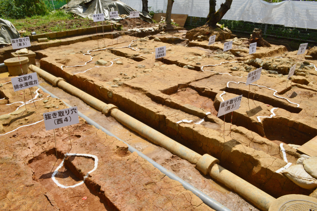 Ein großer Aushub mit Erde, umgeben von Bäumen, Pflanzen, Gras, einem Zaun, einer Abdeckung und einer Wand, mit Schildern, Rohren und verstreuten Gegenständen.