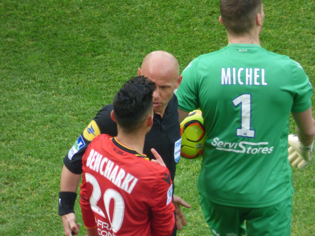 Zwei Fußballspieler und ein Schiedsrichter auf einem grünen Feld, wobei einer der Spieler einen Ball hält.