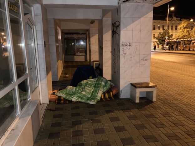 Eine obdachlose Person, die nachts auf einem Bürgersteig schläft, mit einer Decke und einem Hocker in der Nähe, vor einem Gebäude mit einer Tafel, einem Straßenpfosten, Bäumen, Fahrzeugen, Passanten und anderen Gebäuden unter einem bewölkten Himmel.