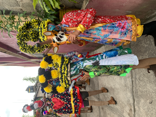 Eine Gruppe von Menschen in bunten Kostümen steht vor einem Gebäude und hält verschiedene Gegenstände, mit Bäumen, Gebäuden und einem klaren blauen Himmel im Hintergrund während eines Faschingsumzugs.