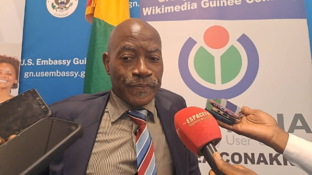 Ein Mann in Anzug und Krawatte spricht mit Reportern, mit einem Mikrofon und einem Aufnahmegerät im Vordergrund und Bannern, Fahnen und einer Frau im Hintergrund.