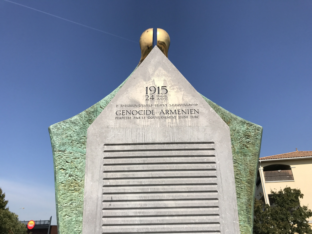 Ein steinernes Denkmal mit einer Uhr darauf steht in einem Park, umgeben von Bäumen und Gebäuden unter einem klaren Himmel, mit einer Inschrift, die den 24. Jahrestag des Völkermords in Armenien gedenkt.