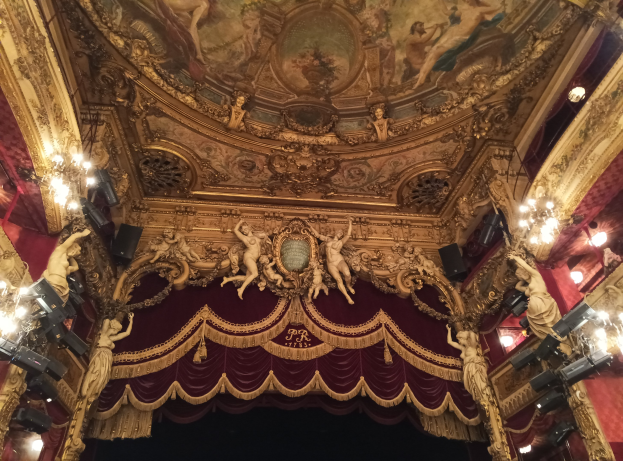 Innere eines Theaters mit einer roten Gardine, Kronleuchtern, Skulpturen, Lichtern, einer Deckenmalerei und einer Bühne mit einigen Menschen davor.