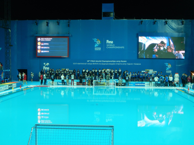 Gruppe von Menschen, die um ein Hallenbad mit Netz am Boden stehen, mit einer Wand, die "FINA World Championships 2015 Kazan"-Text und Logos anzeigt, sowie Trägern und Lampen im Hintergrund.