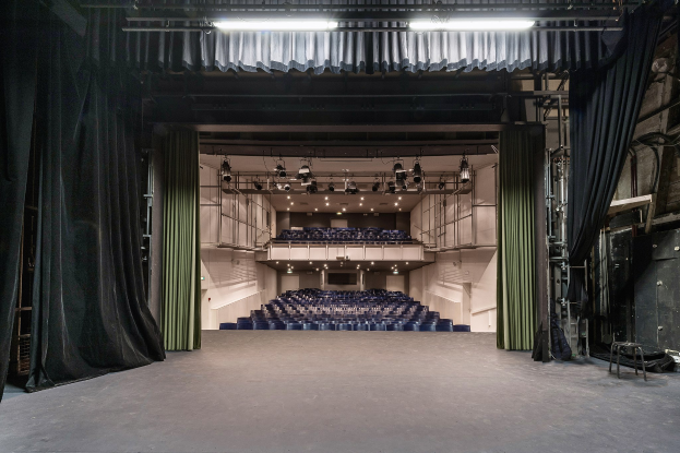 Großer Zuschauerraum mit schwarzen Vorhängen, einer Bühne, Deckenbeleuchtung und für eine Vorstellung angeordneten Stühlen, mit Gegenständen auf der rechten Seite.