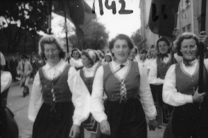 Eine Gruppe von Frauen in traditioneller bayerischer Tracht spaziert straßenabwärts, lächelt und ist von Bäumen und Gebäuden umgeben, auf einem Schwarz-Weiß-Bild.
