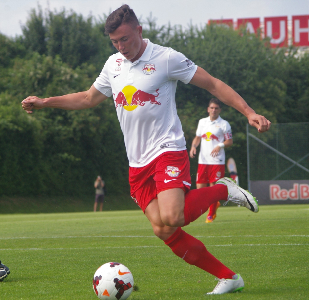 Ein Mann in einem Fußballtrikot kickt einen Ball auf einem grünen Feld mit Bäumen und einem klaren blauen Himmel im Hintergrund, wobei ein Schild mit der Aufschrift "RB Leipzig" sichtbar ist.
