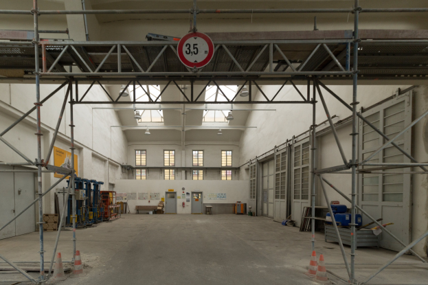 Lagerhalle mit ausgedehnten Gerüsten, Verkehrskegeln, Metallstangen, einem Schild, Maschinen, einer Tür, Fenstern, Wänden, Deckenleuchten und Dach.