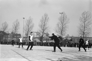 Eine Gruppe von Menschen, die im Schnee Eishockey spielen, mit Bäumen, Gebäuden, Laternenmasten und einem klaren Himmel im Hintergrund, dargestellt in Schwarz-Weiß.