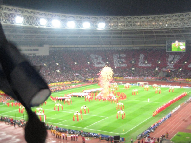 Ein Stadion mit Flutlicht, Eisengittern, Zuschauern, stehenden Personen, Werbetafeln, Bildschirmen, einer Kamera und Fahnen und Dekorationen.