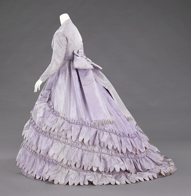 Eine Schaufensterpuppe in einem lila Ballkleid mit Rüschen an den langen Ärmeln, einer eng anliegenden Taille und einem bodenlangen Rock vor einem grauen Hintergrund.