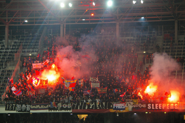 Große Menschenmenge in einem Stadion mit Feuerwerkskörpern und Rauch, haltend Fahnen und Banner, unter einem Dach mit Deckenbeleuchtung.